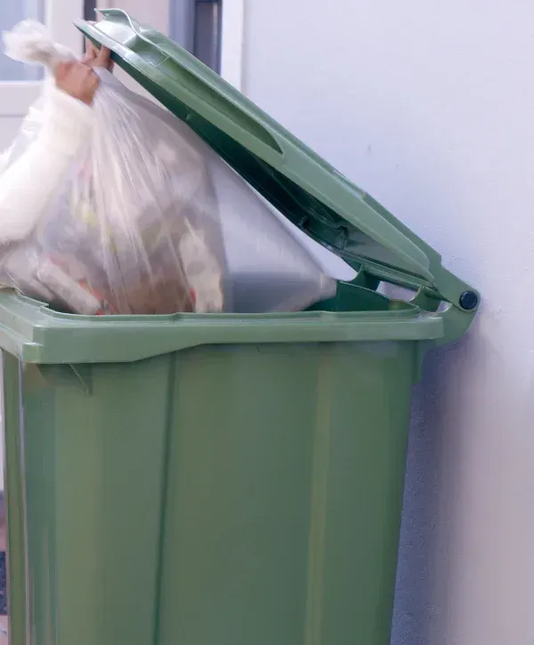 A person's hands placing a large clear plastic bag full of trash into a dark green outdoor waste bin with its lid open.