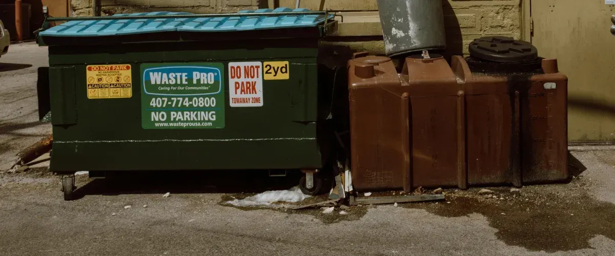 A green Waste Pro dumpster with blue lid and brown trash cans alongside a wall on a paved surface.