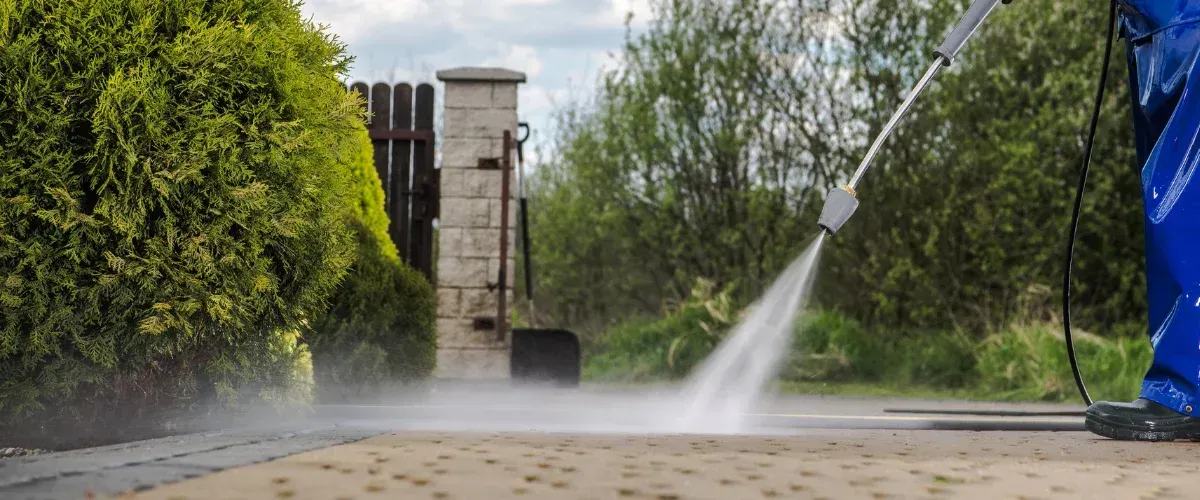 A person in blue overalls power-washing a paved driveway next to green bushes and a stone gate.