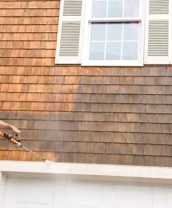 A man in a blue hard hat and safety glasses power washes a red tiled roof with a skylight on a sunny day.
