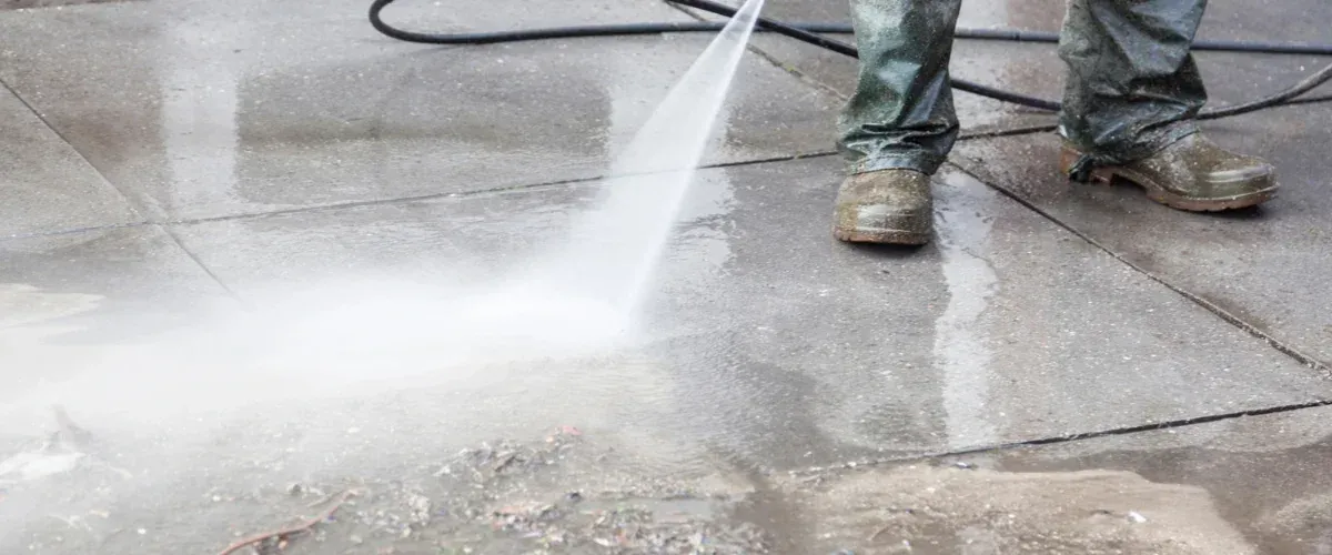 A person in olive green waterproof pants and boots pressure washing a gray concrete sidewalk, with water and dirt spraying.