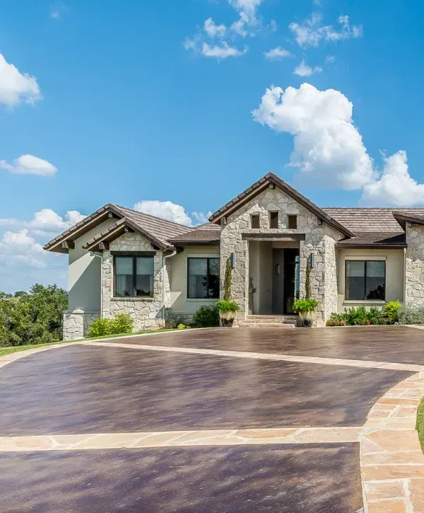 A luxurious modern stucco and stone house with a circular, patterned dark concrete driveway under a bright blue sky.