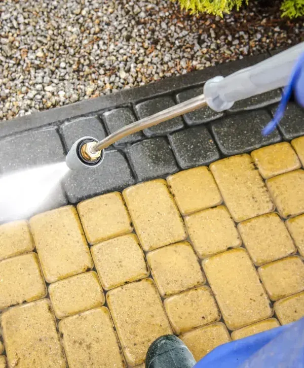 A person uses a pressure washer to clean dirty yellow patio pavers, revealing clean, wet black pavers beyond.