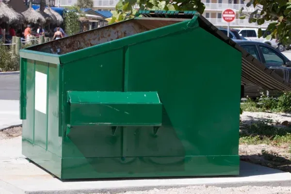 A large, weathered green dumpster sits on a concrete slab outdoors, with a beachside resort visible in the blurred background.