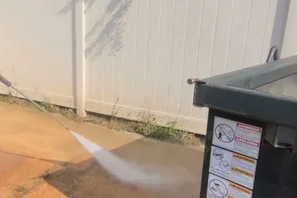 A person pressure washing dirty ground next to a green dumpster and a white fence on a sunny day.