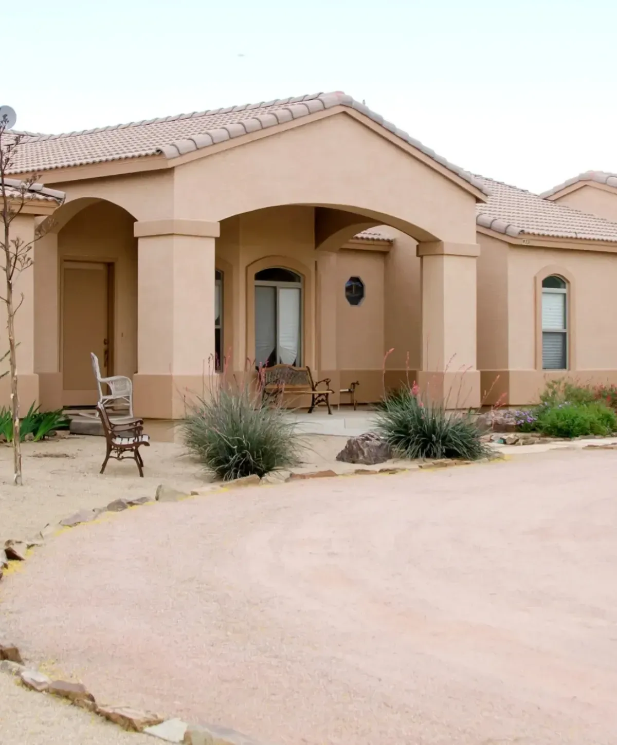 A tan stucco home with an arched porch entryway and tiled roof, featuring desert landscaping and a gravel driveway.
