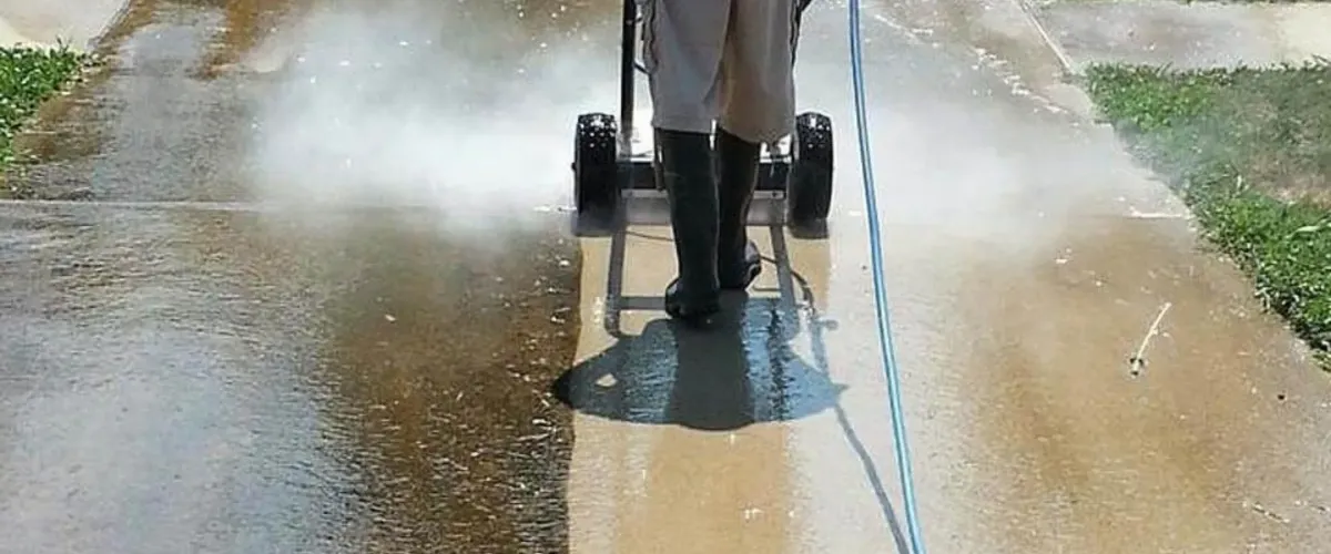 A person uses a pressure washer surface cleaner on a dirty driveway, showing a clear line between cleaned and uncleaned concrete.