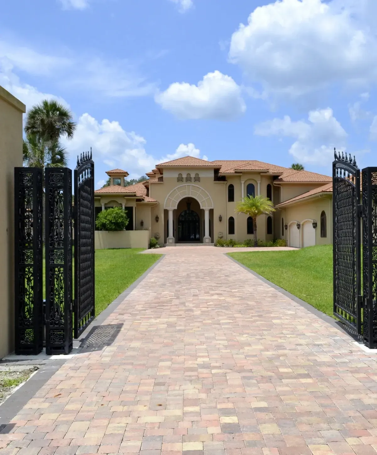 A grand Mediterranean-style estate with a tan stucco exterior and tiled roof, seen through open ornate black iron gates and a brick paver driveway.