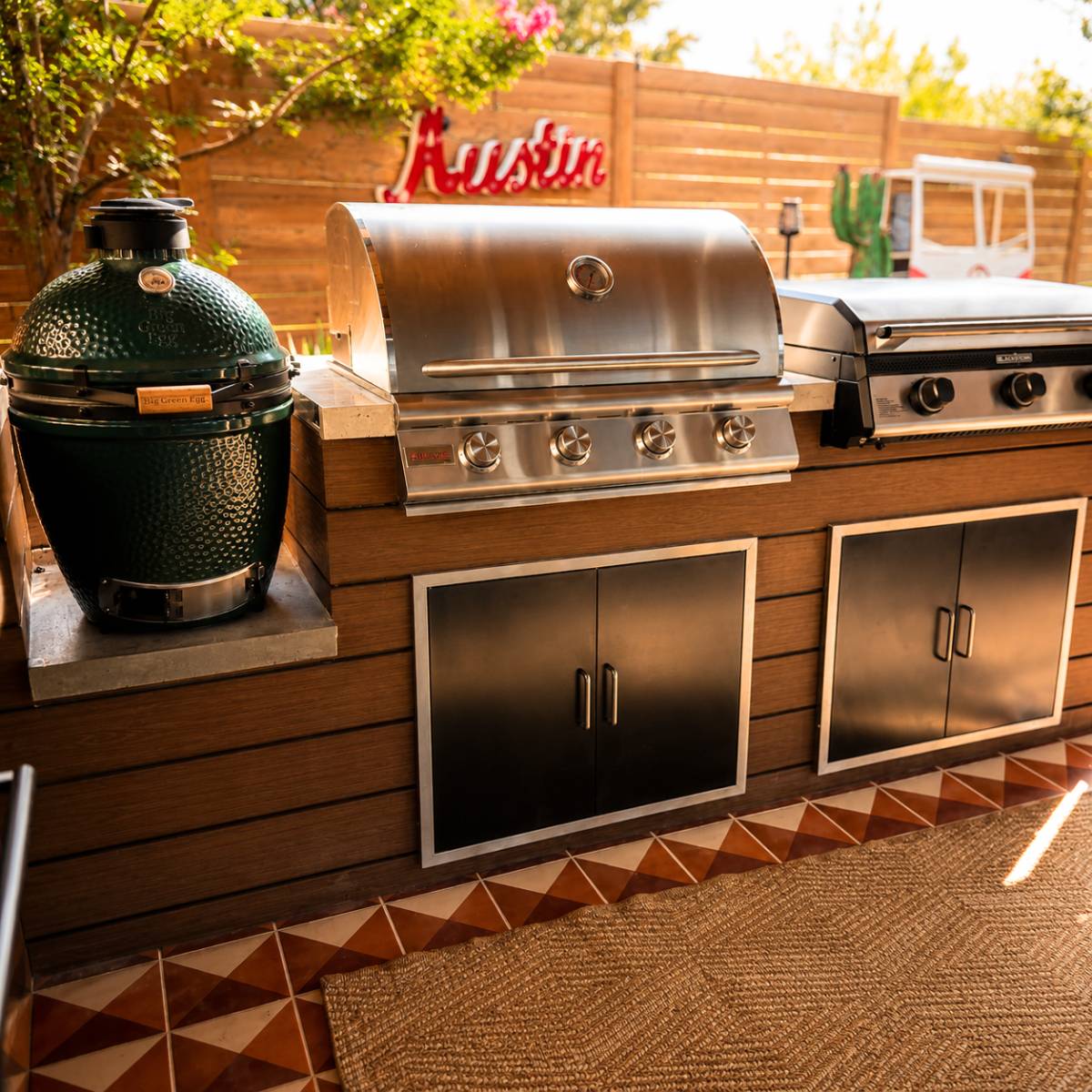 Custom outdoor kitchen remodel in Austin, Texas featuring a built-in stainless steel gas grill, Big Green Egg smoker, and Blackstone griddle integrated into a modern wood-clad island with stainless steel storage cabinets.