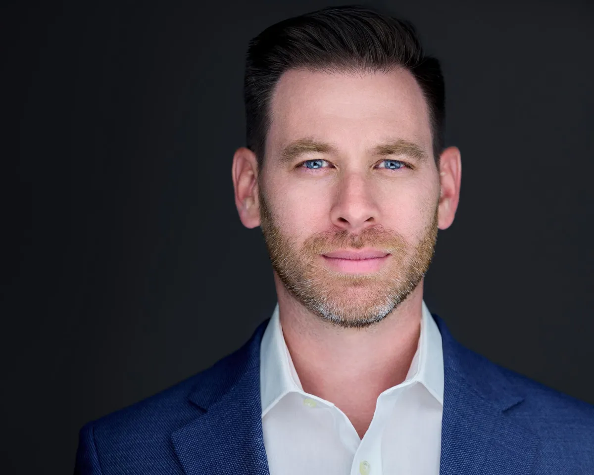 Professional headshot of a man in a blue suit and white shirt, looking confident against a dark background