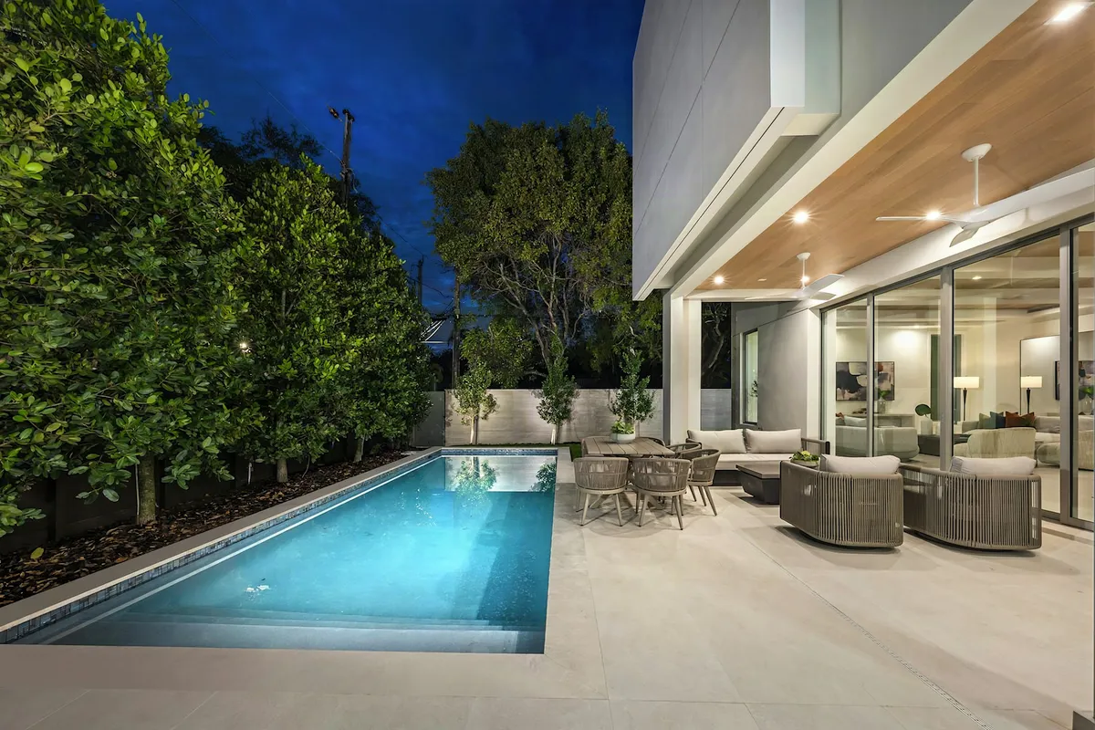 Lit concrete pool beside a modern home and outdoor living area at dusk