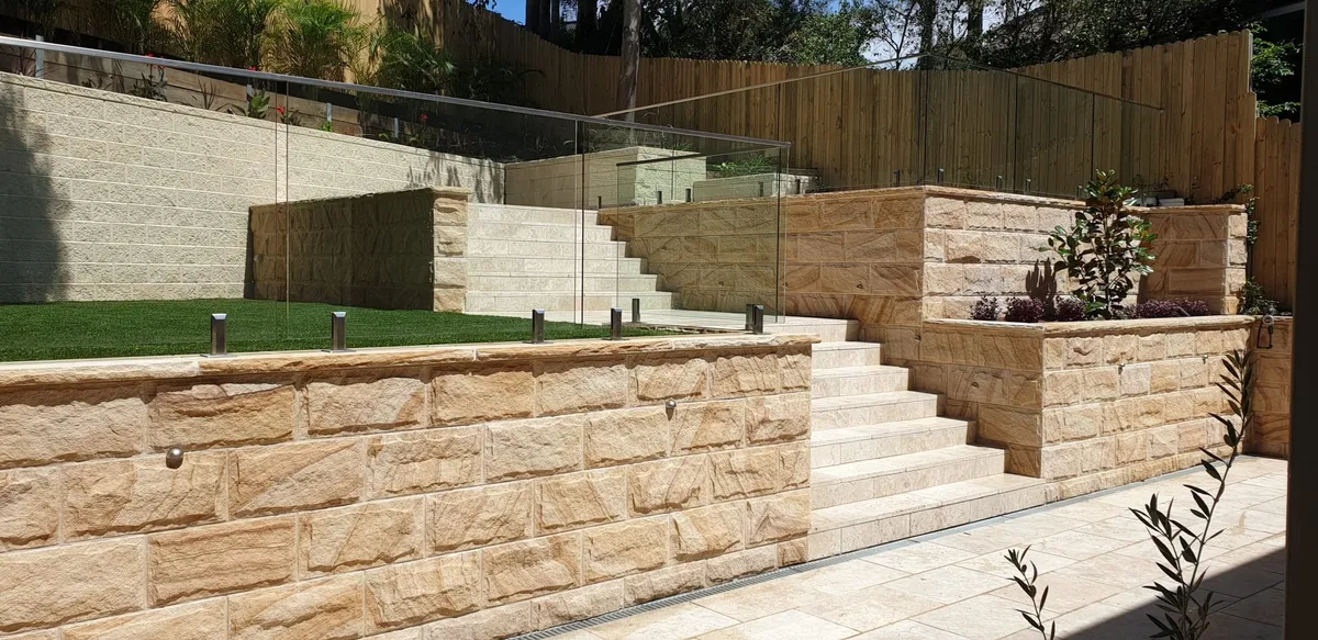 Terraced sandstone retaining walls with stone stairs, glass fencing and travertine paving