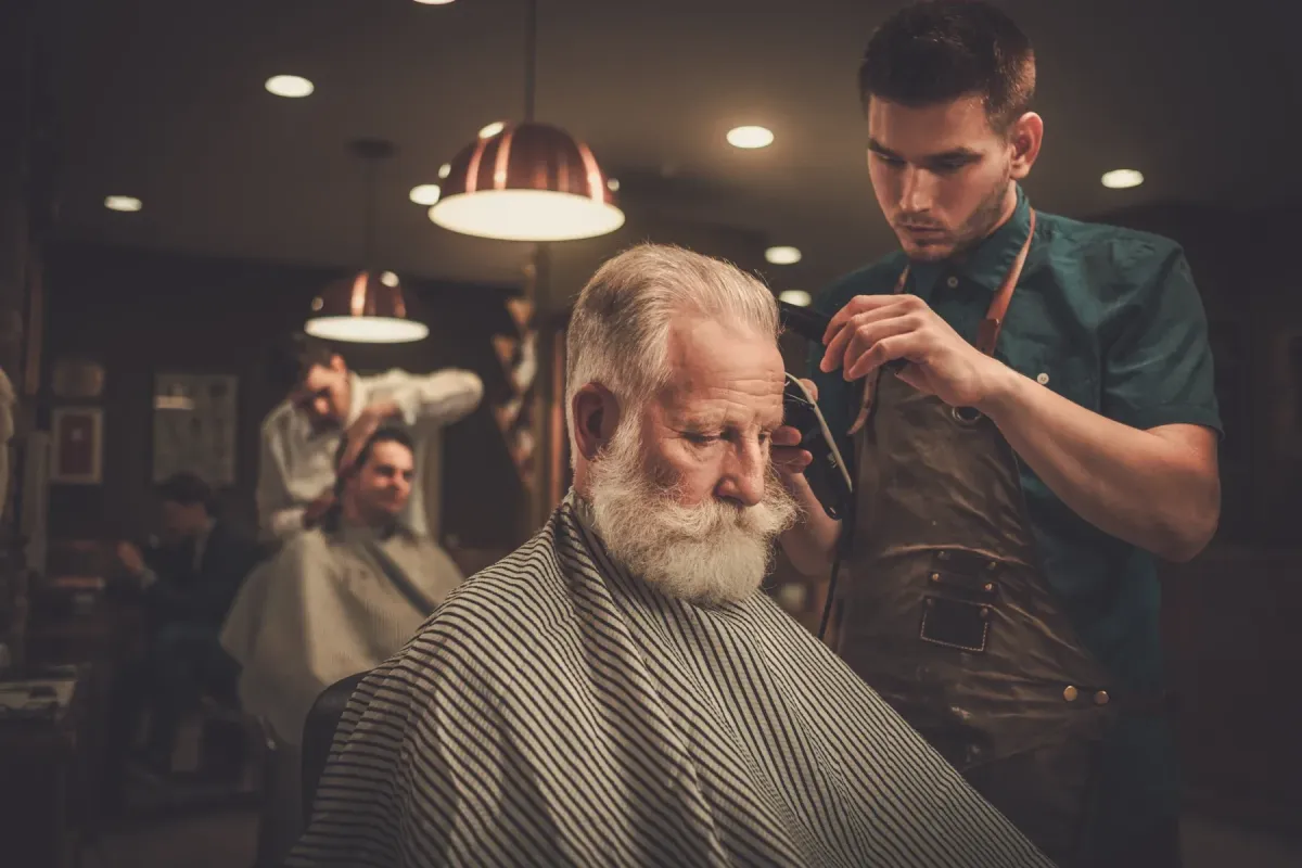 Barber at Scuttlebutt Barbershop in Portsmouth, NH trimming an older gentleman’s beard in a warmly lit, vintage-style barbershop.
