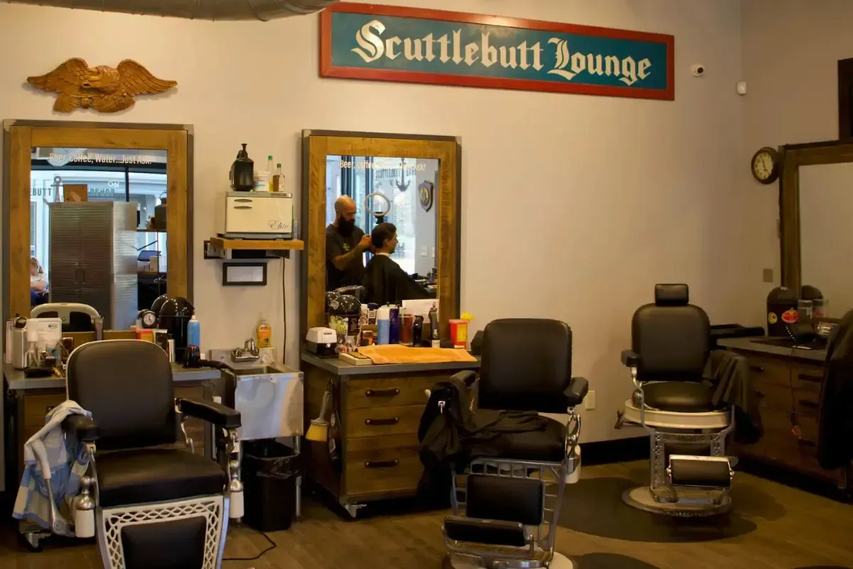 Interior of Scuttlebutt Barbershop in Portsmouth, NH featuring vintage barber chairs, wood-framed mirrors, and a wall sign that reads 'Scuttlebutt Lounge'