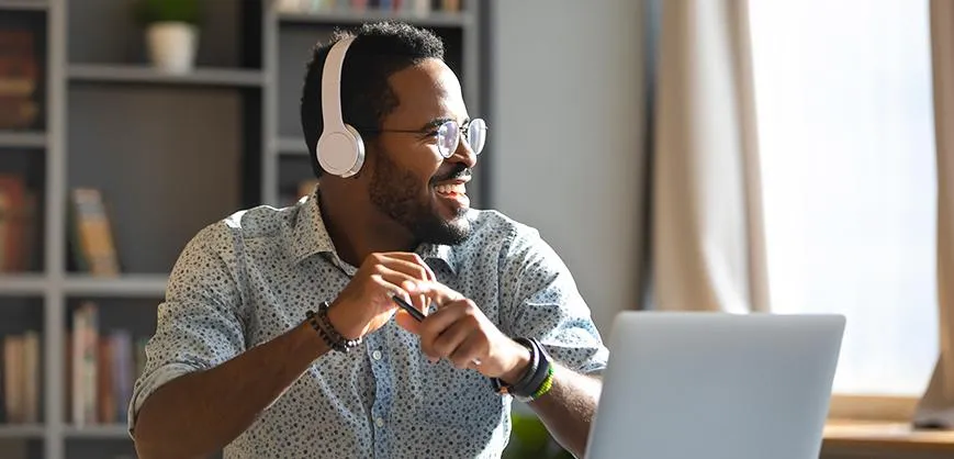 Smiling man wearing headphones, engaged in an online meeting or virtual collaboration from a home office. He is sitting at a desk with a laptop, appearing enthusiastic and focused. The background features bookshelves and natural lighting, highlighting a comfortable remote work environment.