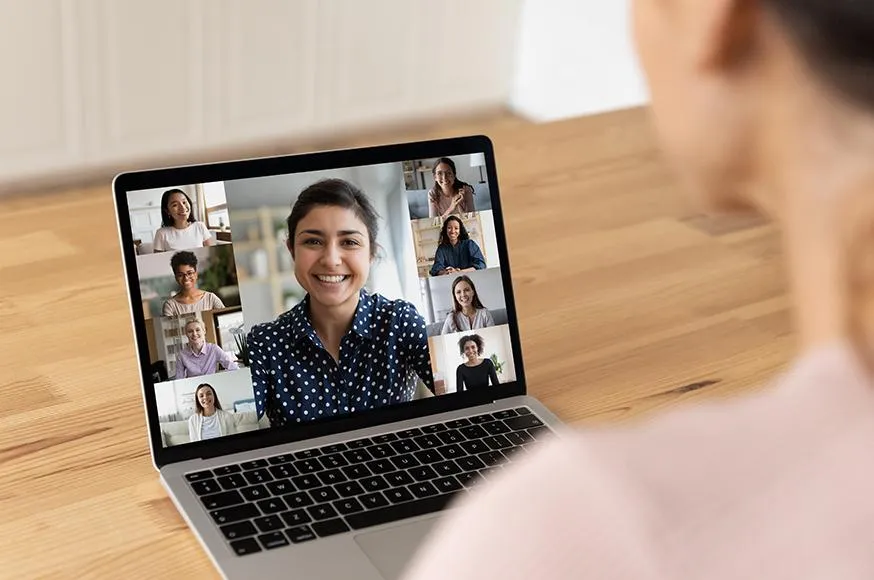 Person participating in a virtual video conference call on a laptop. The screen displays a diverse group of smiling professionals engaged in an online meeting. The setting is a well-lit, modern home office, emphasizing remote work, virtual collaboration, and online communication.