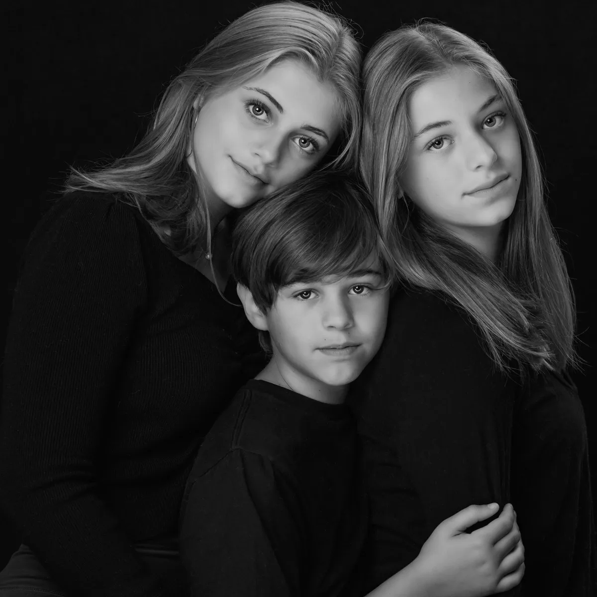 Three siblings lean close together in a black-and-white portrait, their heads gently touching as they look calmly toward the camera (Evidence), sharing a quiet sense of trust, closeness, and ease with one another (Emotion), reflecting the deep bond and belonging that grows from being shaped side by side over time (Elevation).