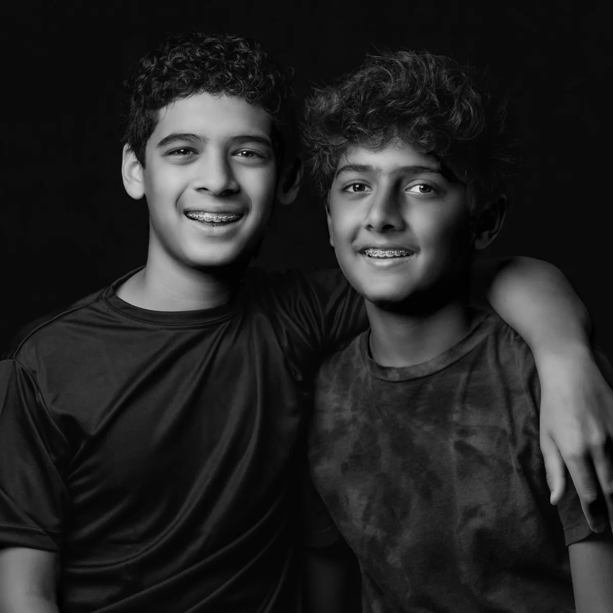 Two siblings stand close together in a black-and-white portrait, one with an arm around the other as they smile gently toward the camera (Evidence), sharing a feeling of ease, trust, and quiet confidence with one another (Emotion), reflecting the steady bond and belonging that comes from growing up side by side and knowing you’re not alone (Elevation).