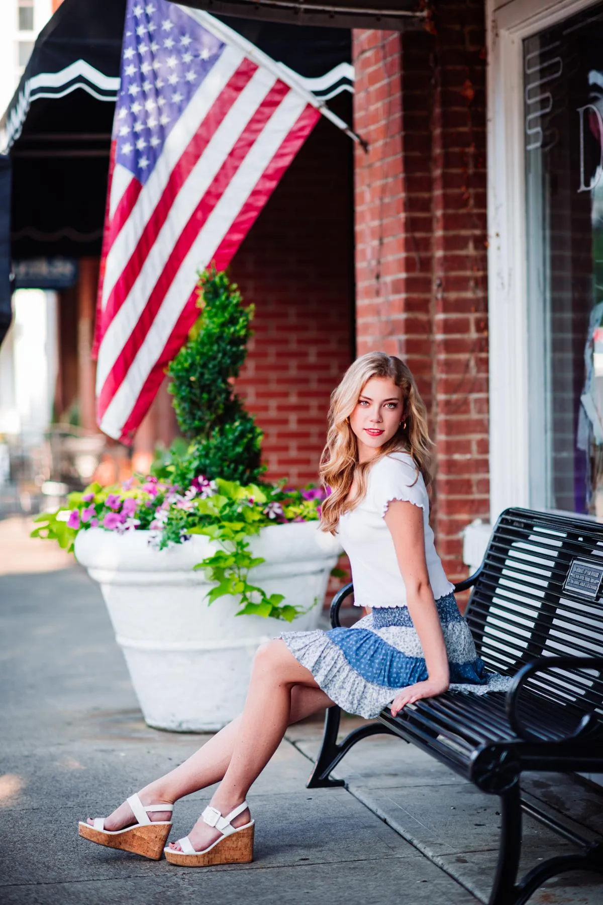 A young woman sits confidently on a bench along a brick-lined sidewalk, with an American flag and storefronts behind her as she looks toward the camera (Evidence), feeling self-assured, poised, and ready for what’s next (Emotion), reflecting the confidence that comes from stepping into her own voice and direction in life (Elevation).