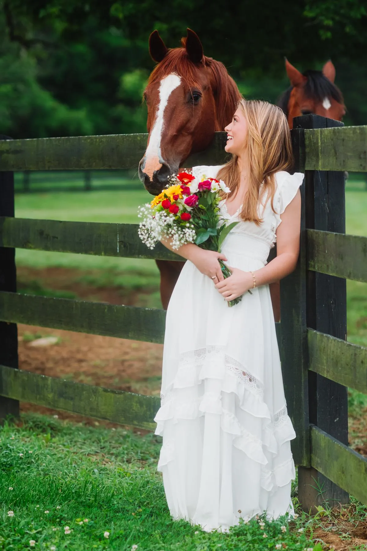 A young woman in a flowing white dress stands by a wooden fence holding a bouquet of colorful flowers, smiling toward a horse beside her in a green outdoor setting (Evidence), feeling joyful, present, and gently connected to the moment around her (Emotion), reflecting a sense of harmony, confidence, and appreciation for the life she is living right now (Elevation).