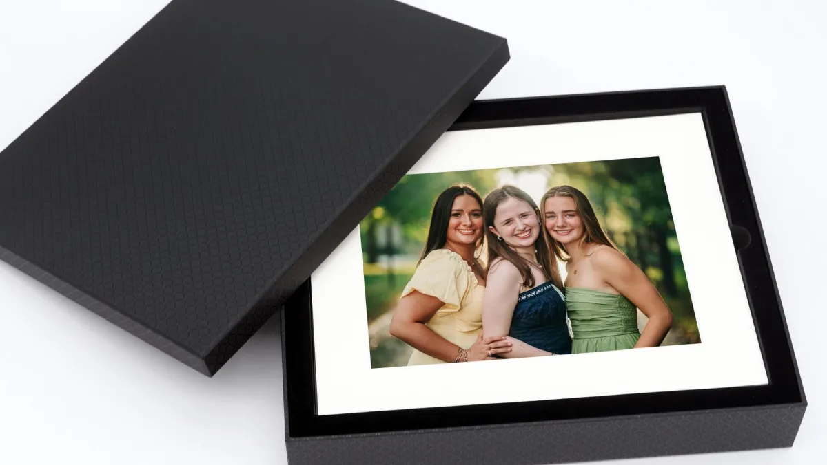 A framed portrait of three young women rests inside an open, textured keepsake box, carefully presented on a clean surface (Evidence), carrying a feeling of pride, warmth, and togetherness (Emotion), reflecting how shared relationships are honored and preserved as part of a family’s story and home (Elevation).