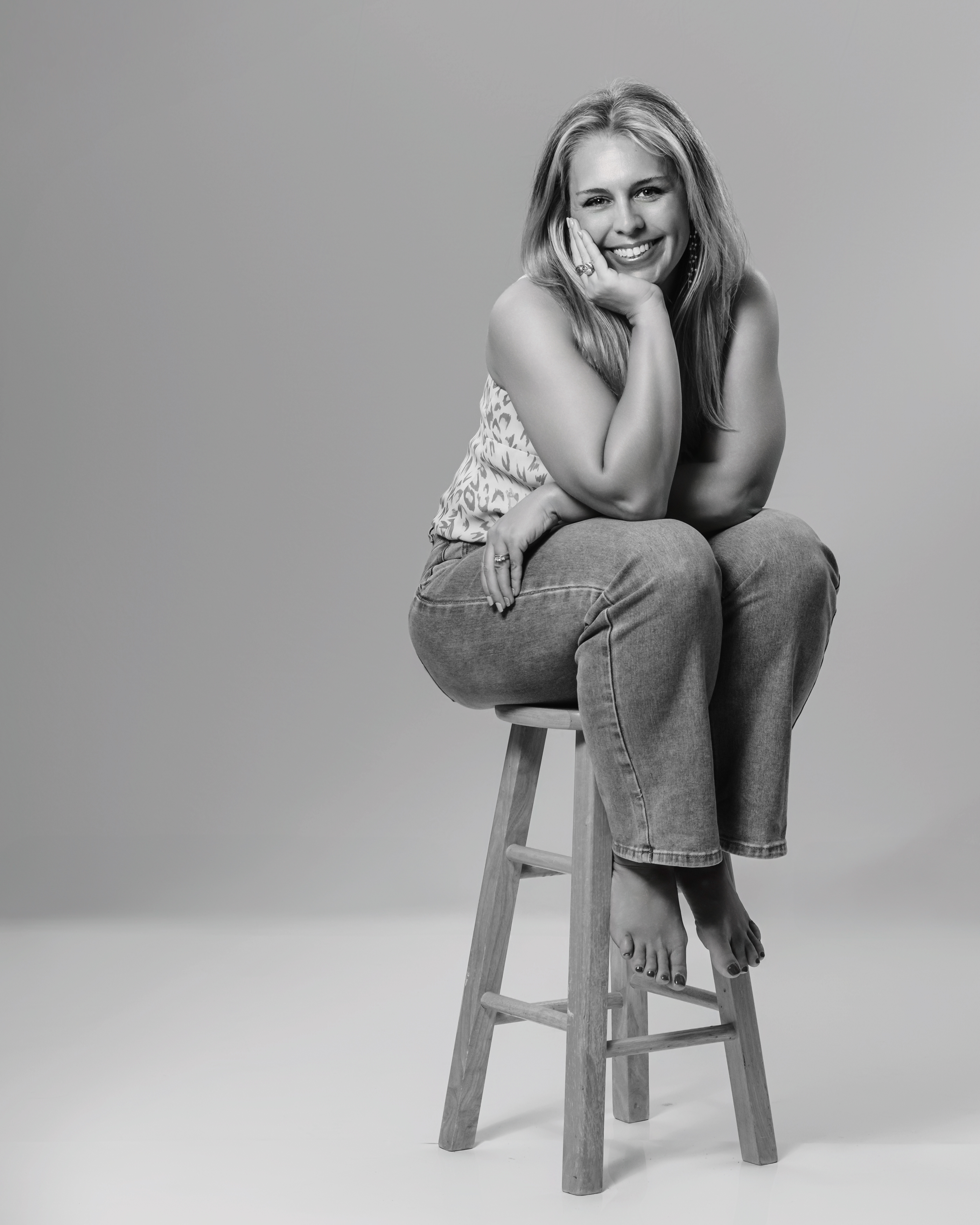 A woman sits barefoot on a wooden stool in a simple studio space, resting her chin in her hand and smiling gently toward the camera (Evidence), feeling relaxed, open, and quietly confident (Emotion), reflecting the ease that comes from being fully seen and comfortable in who she is (Elevation).