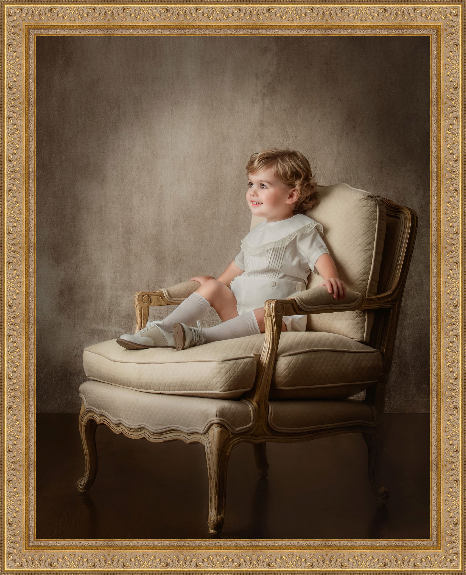 A young child sits comfortably in an upholstered armchair, dressed in light clothing and looking off to the side with a gentle smile, framed by an ornate border (Evidence), feeling calm, curious, and quietly content (Emotion), reflecting the innocence and presence that families treasure and choose to honor as part of their home and story (Elevation).