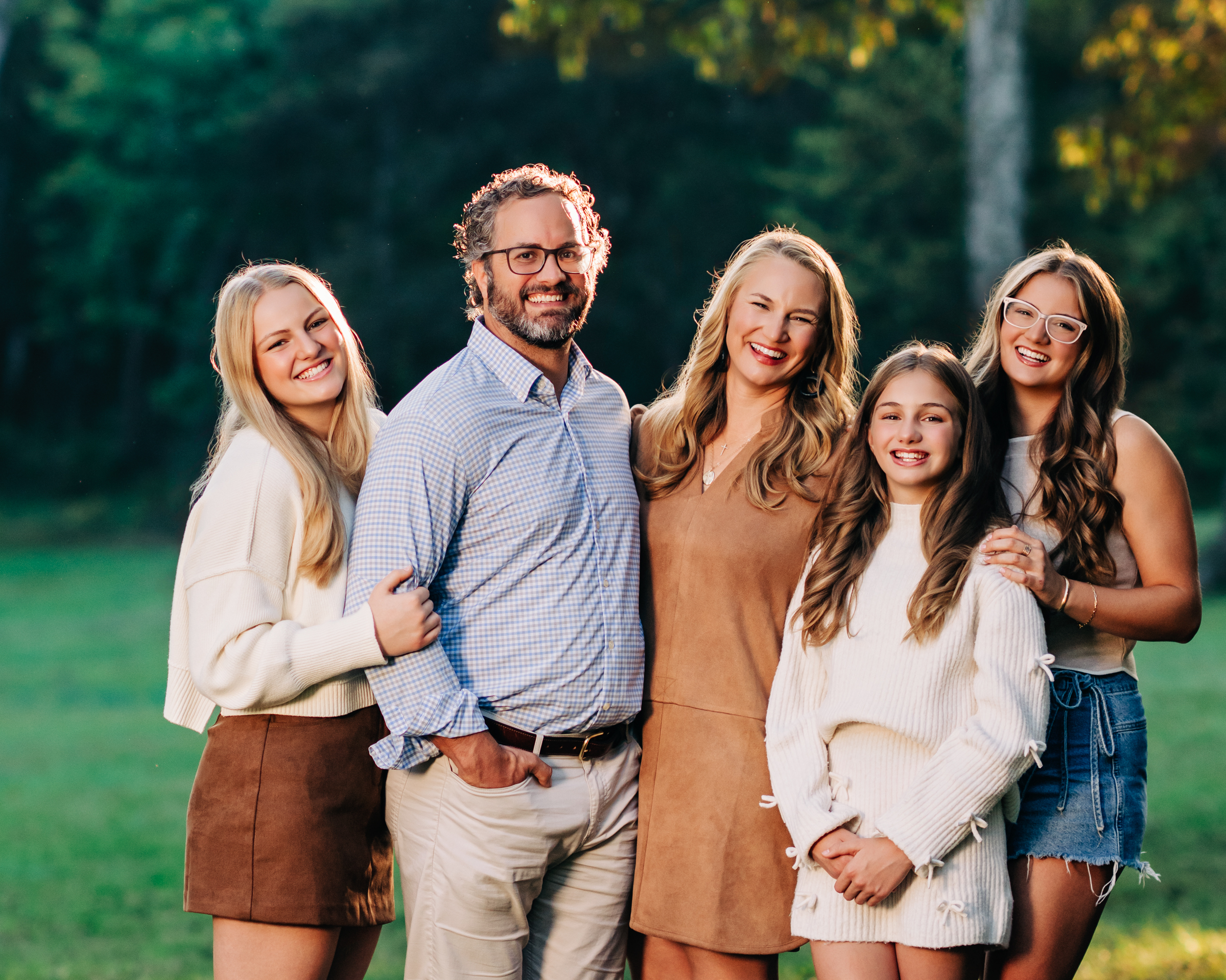A family of five stands close together outdoors, arms linked and leaning into one another while smiling naturally (Evidence), sharing a feeling of warmth, ease, and genuine connection (Emotion), reflecting the security and belonging that comes from being known and supported within their family (Elevation).