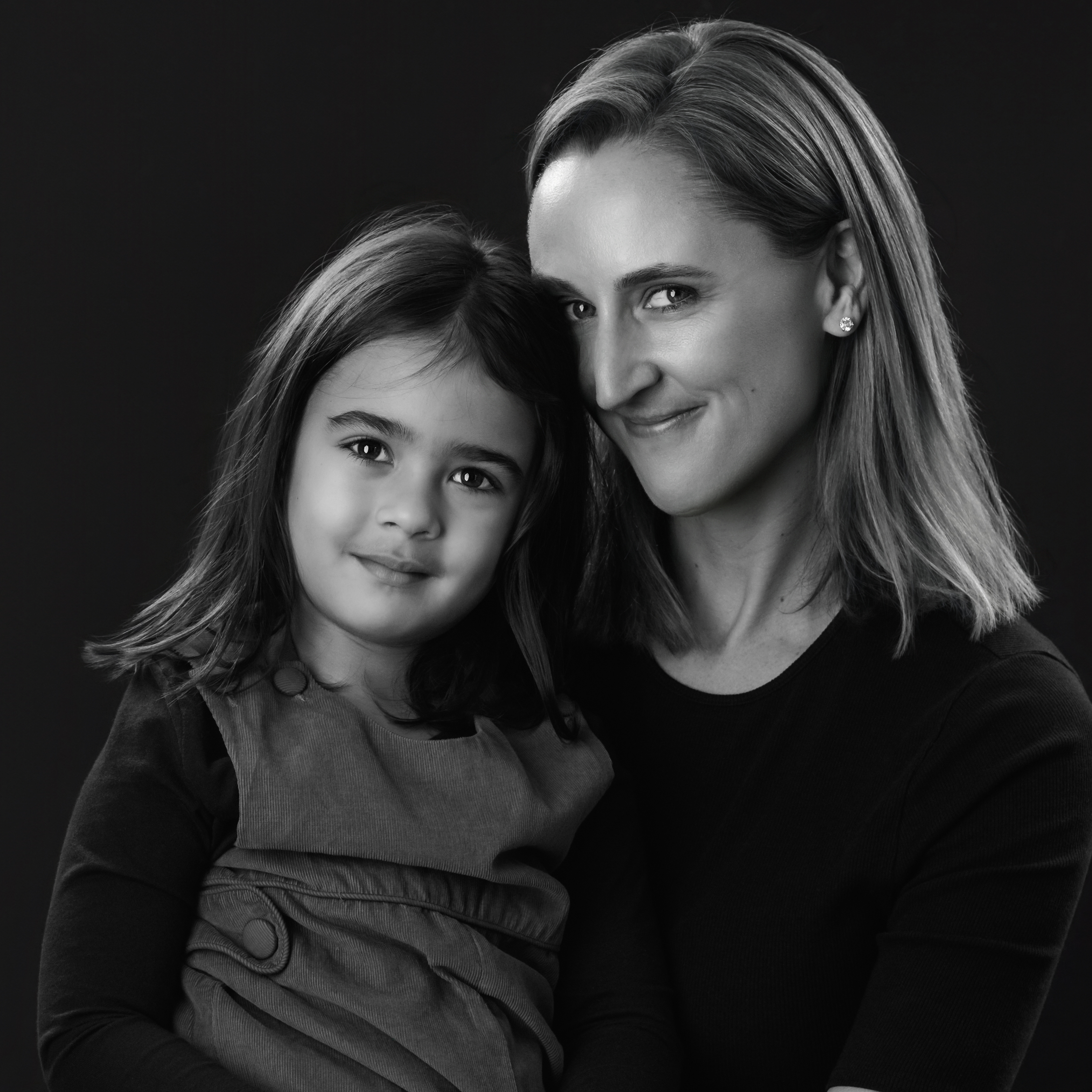 A woman and a young child sit close together in a black-and-white portrait, their faces touching as they look calmly toward the camera (Evidence), sharing a feeling of tenderness, trust, and quiet closeness (Emotion), reflecting the deep bond and sense of safety that grows from being lovingly held and known (Elevation).