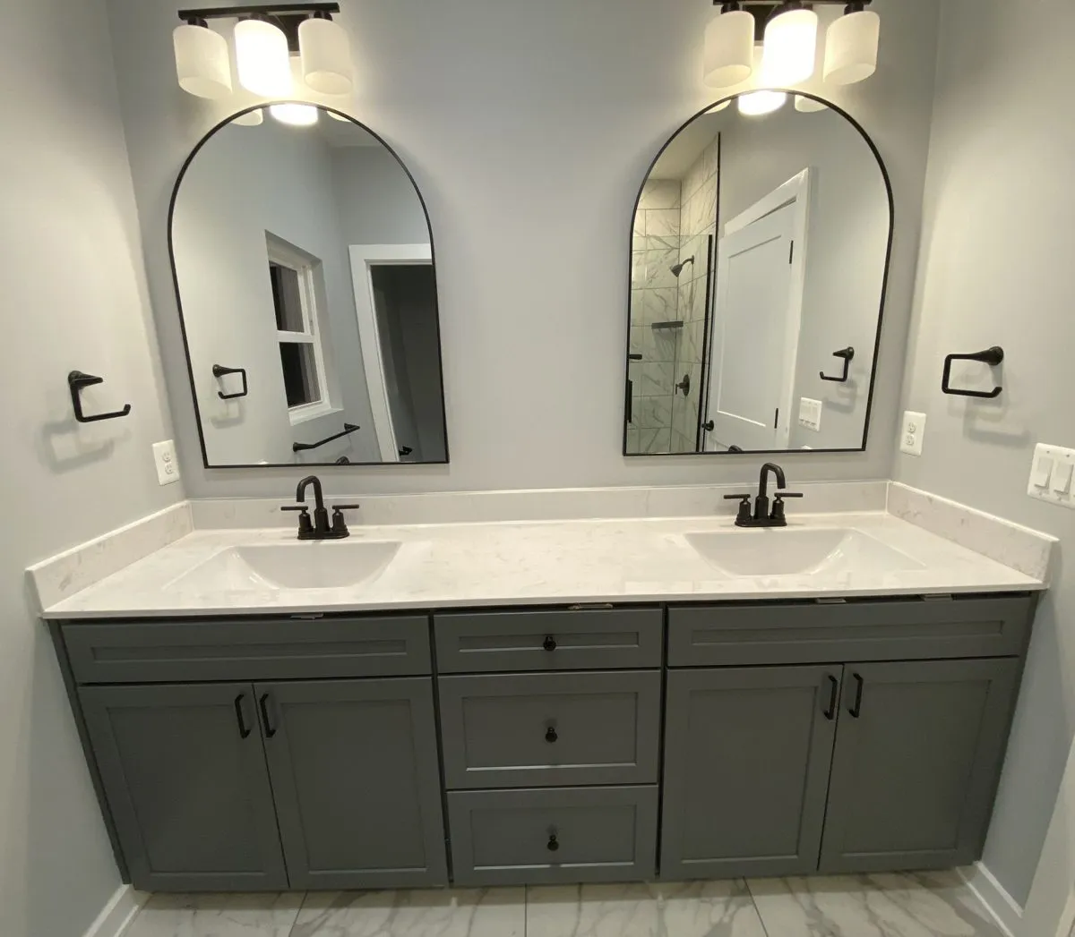 Double vanity with gray shaker cabinets, two sinks, matte black faucets and arched black-framed mirrors