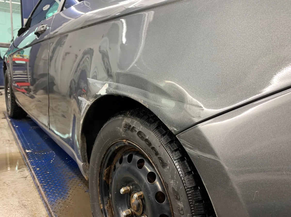 Voiture grise avec bosse visible sur l’aile arrière gauche et portière, en cours de nettoyage avant réparation.