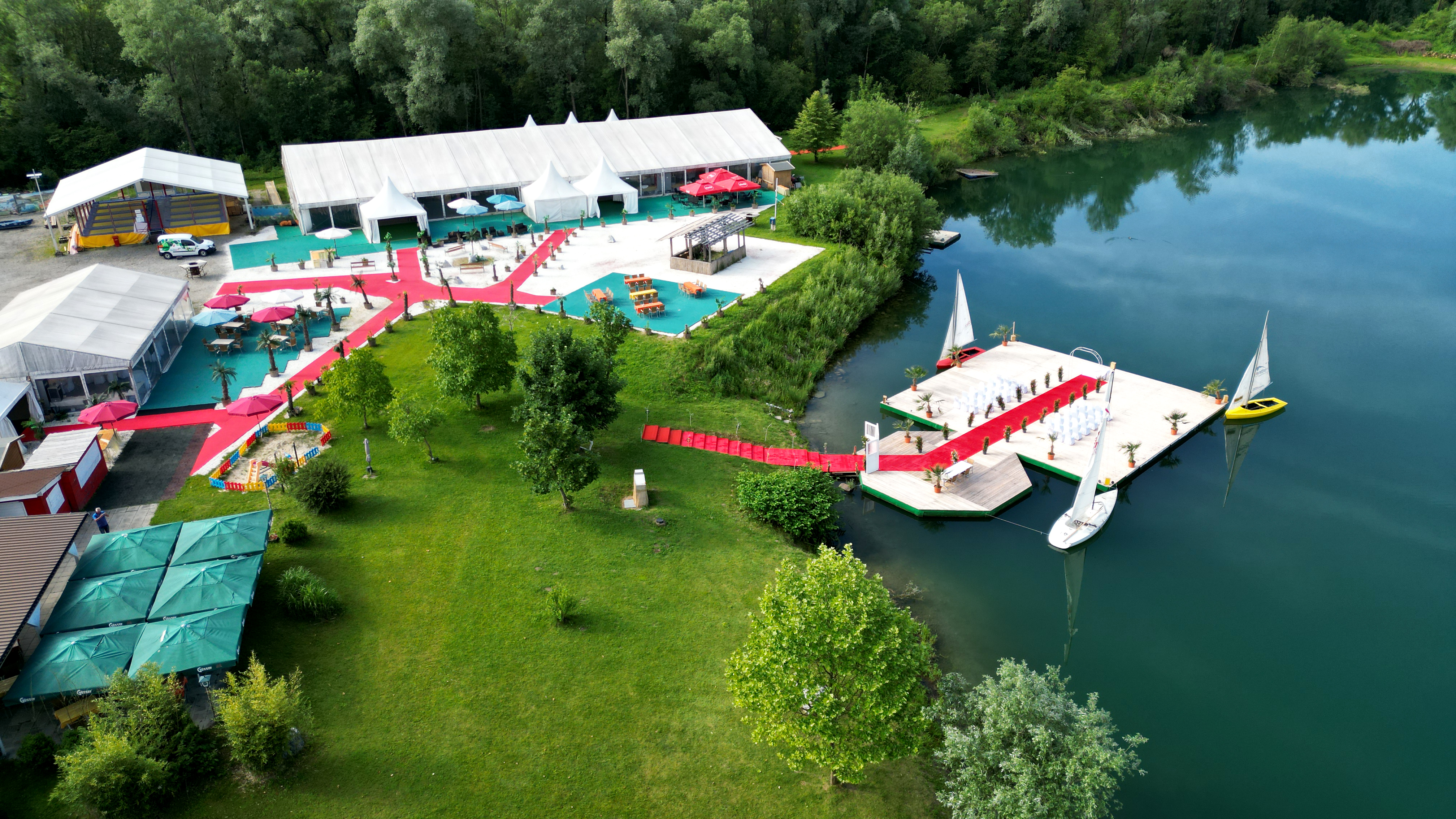 Eventsteg am Badesee bei Flasch City near Suedsteiermark mit rotem Weg und Gesamtuebersicht der Anlage, umgeben von Gruenflaechen und Wasser