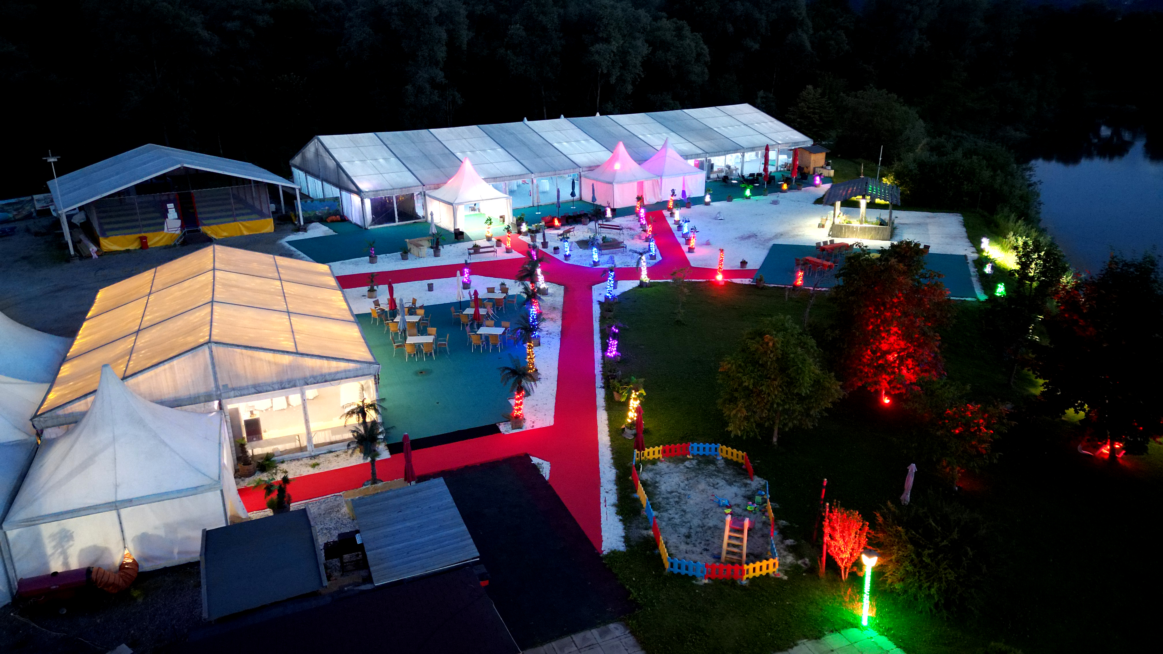 Nacht Luftaufnahme von Flasch City highway stop near mueller-kapfenberg mit beleuchteten Zelten, roten Wegen und Rastplatz am See
