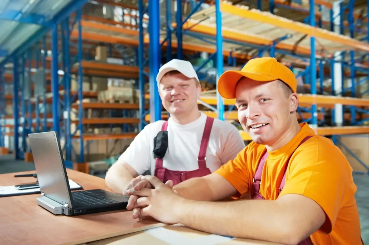 Two smiling warehouse workers sitting at a desk with a laptop.