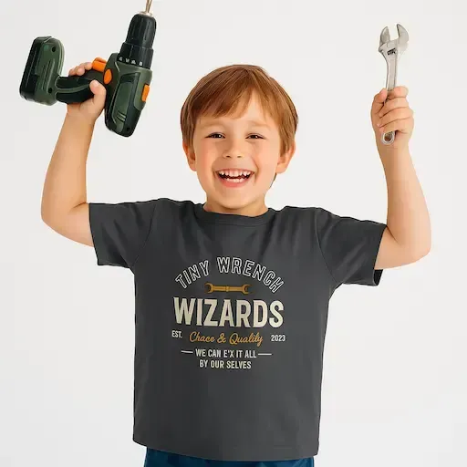 A smiling child holds a drill and a wrench, wearing a dark T-shirt with text. Highlighting toddler clothing and original designs.