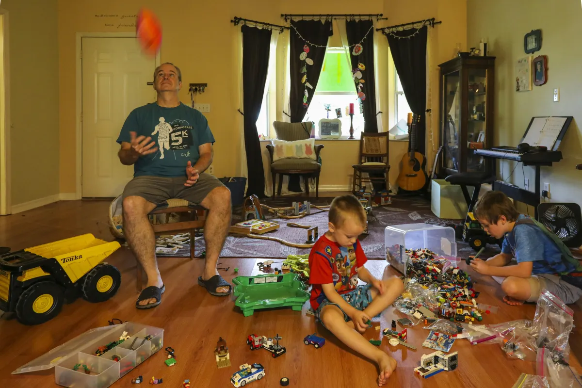 Living room filled with toys, 2 kids playing on the floor, adult nearby in a chair throwing a ball in the air. A great example of parallel playing