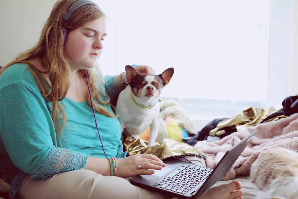 Young female-presenting individual sitting cross-legged on bed, wearing headphones on computer with support dog