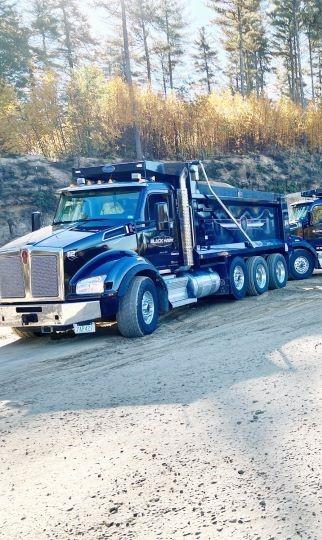 Black Hawk Paving black dump truck parked on a construction site with a scenic forest backdrop.