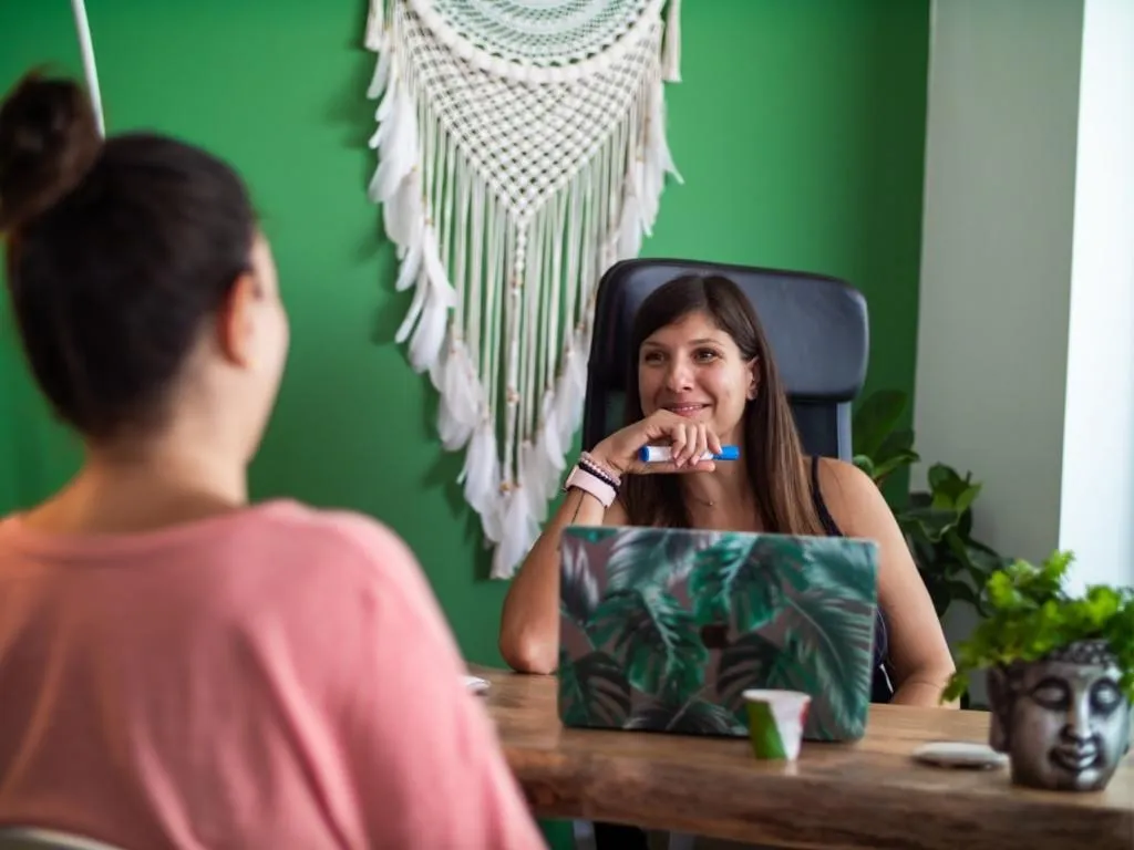 dark haired woman sitting at desk with laptop in front of her smiling talking to a woman in the foreground