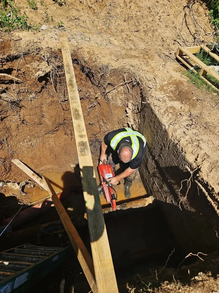 Worker in a safety vest repairing an underground pipe inside a deep trench at a utility excavation site.