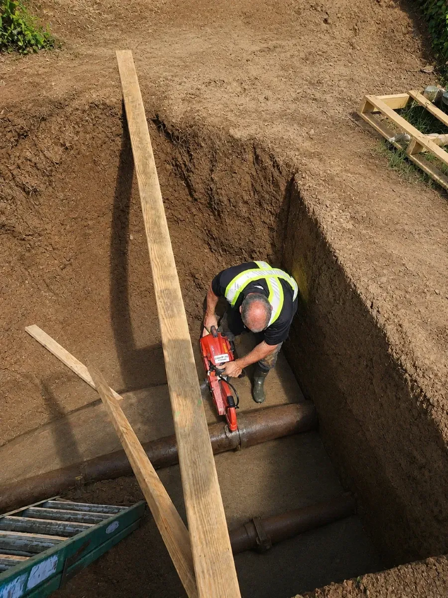Worker in a safety vest repairing an underground pipe inside a deep trench at a utility excavation site.