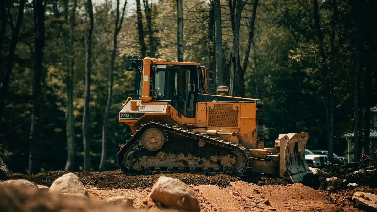 Tracked bulldozer with its blade lowered beside trees and exposed soil.