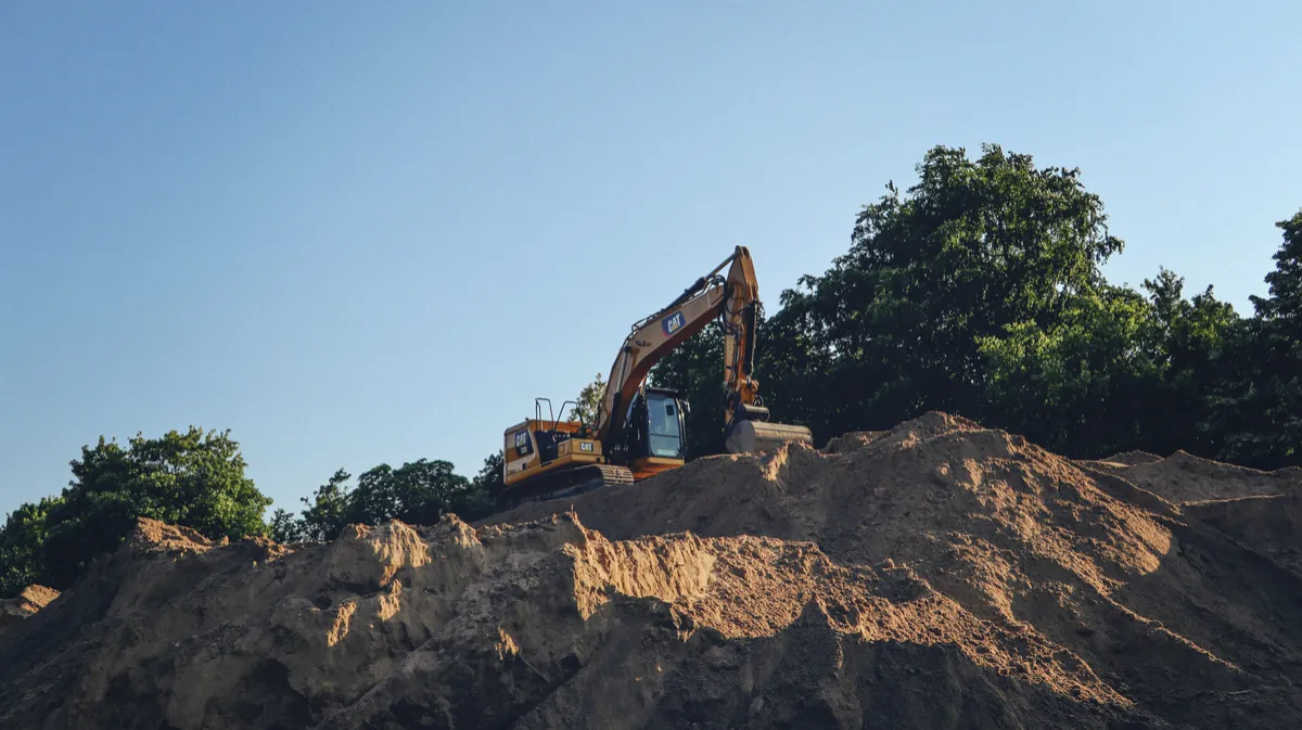 Tracked excavator above an exposed earth cut beside trees.