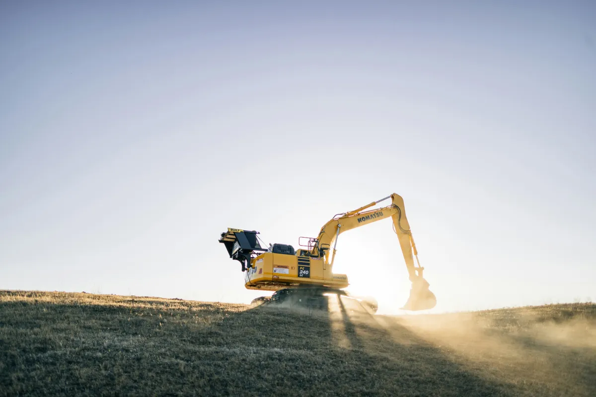 Tracked excavator on a grassy earthwork site in low sunlight.