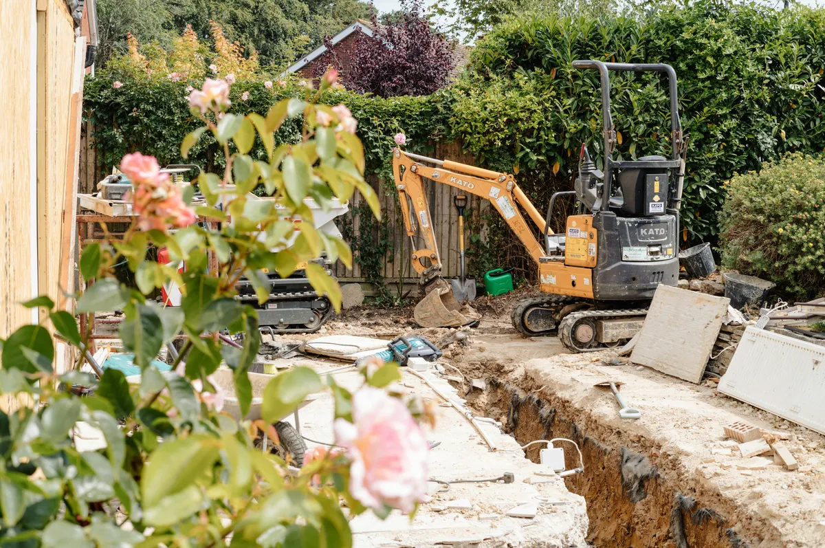 Compact excavator beside a narrow residential earthwork trench.