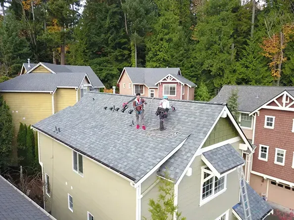 Roofing contractors working on a residential roof in a suburban neighborhood, using safety harnesses and equipment while standing on asphalt shingles surrounded by trees and nearby homes.