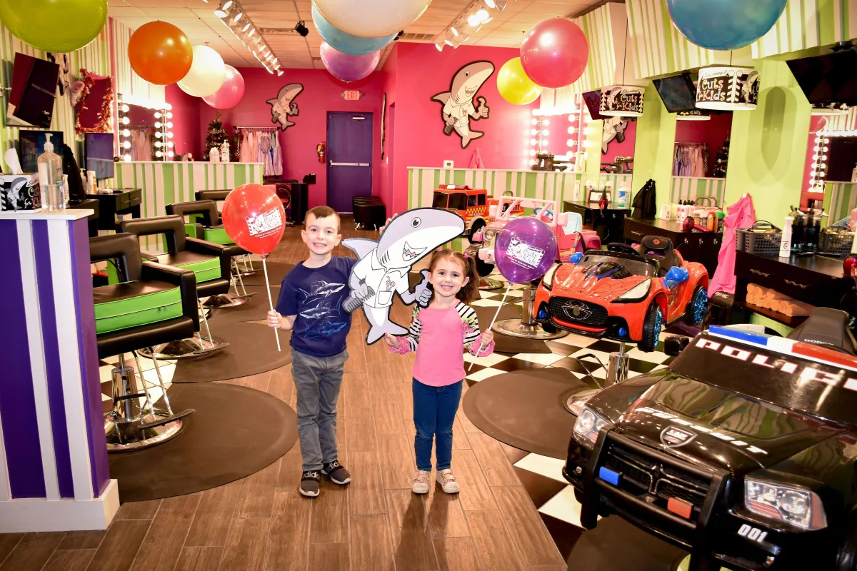 Two children smiling and holding balloons inside Sharkey's Cuts for Kids in Short Pump, surrounded by colorful hair-cutting stations designed like cars, under a festive atmosphere with balloons and cartoon shark decor.