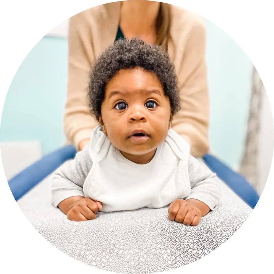A baby with curly black hair rests on a patterned pillow, being adjusted by Dr Nikki in a beige cardigan, in a soothing environment.