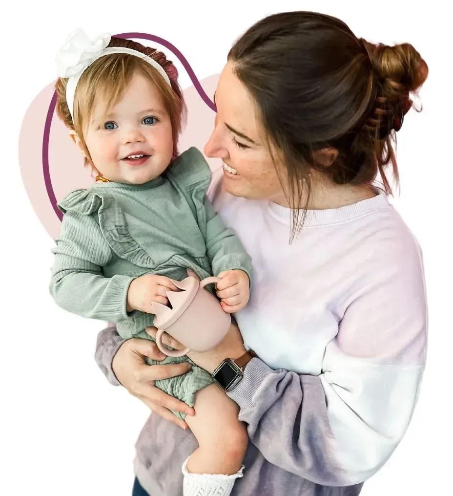 A smiling woman with her hair in a messy bun holds a happy baby wearing a white headband and a green outfit. The baby is holding a pink sippy cup, and the woman is wearing a tie-dye sweatshirt and a smartwatch. A purple heart-shaped outline frames the scene.
