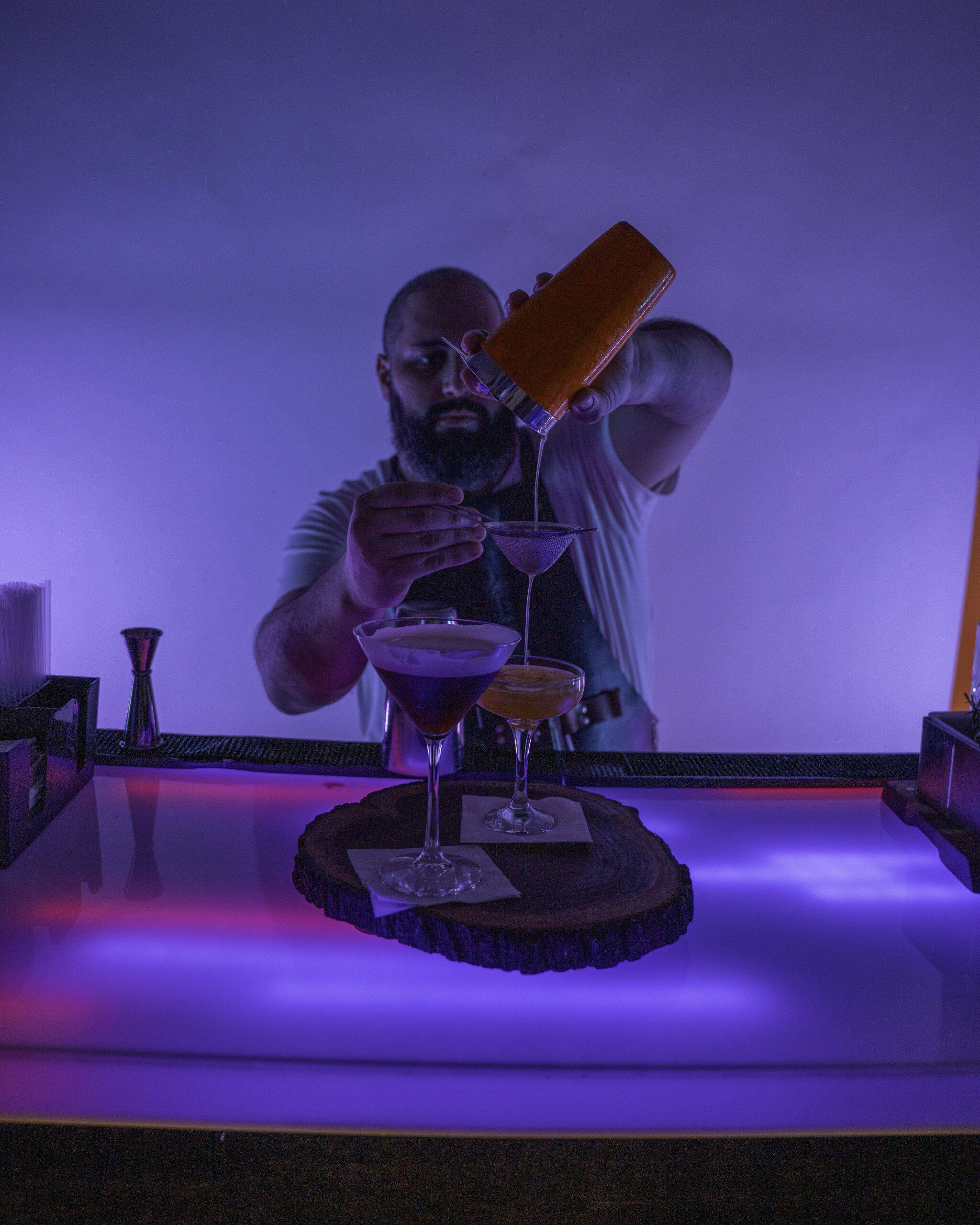 Bartender pouring a cocktail into a glass behind a bar.